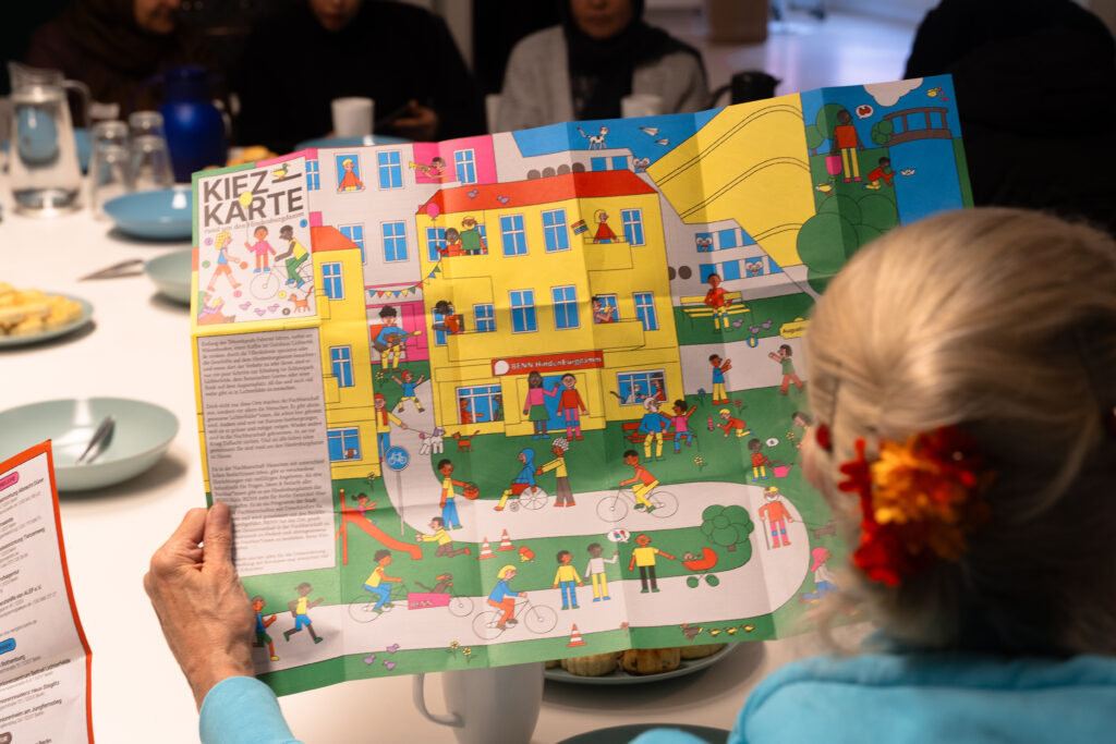 An einem Tisch sitzen viele Menschen. Davon hält eine Frau eine große faltbare Karte in der Hand.