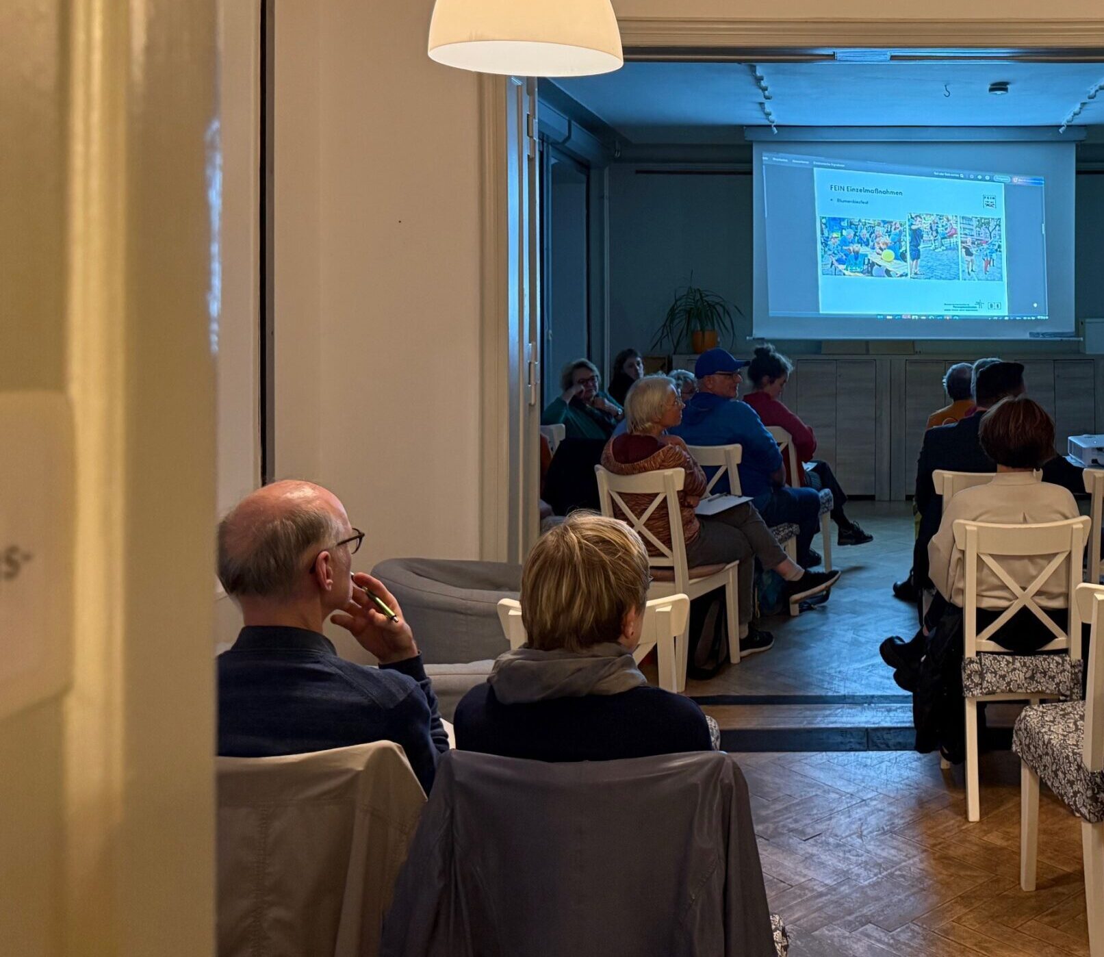 In einem großen Raum sitzen Menschen in Reihen auf weißen Stühlen. Davor wird mit Beamer eine Präsentation gezeigt.